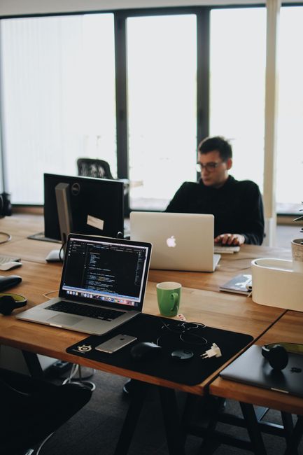 An office space with a large wooden table positioned in front of floor-to-ceiling windows that let in natural daylight. On the table, there are multiple laptops, including a silver one with an open co