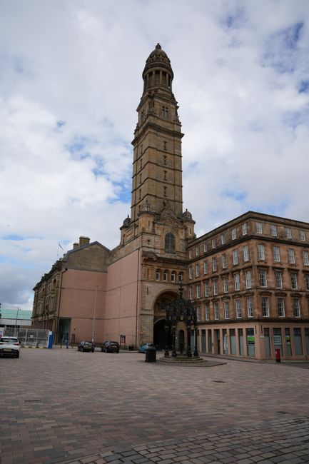 The image depicts a historic building with large, ornate stone columns and decorative architectural details, situated in an urban setting. In front of the building, a bronze equestrian statue of a mou